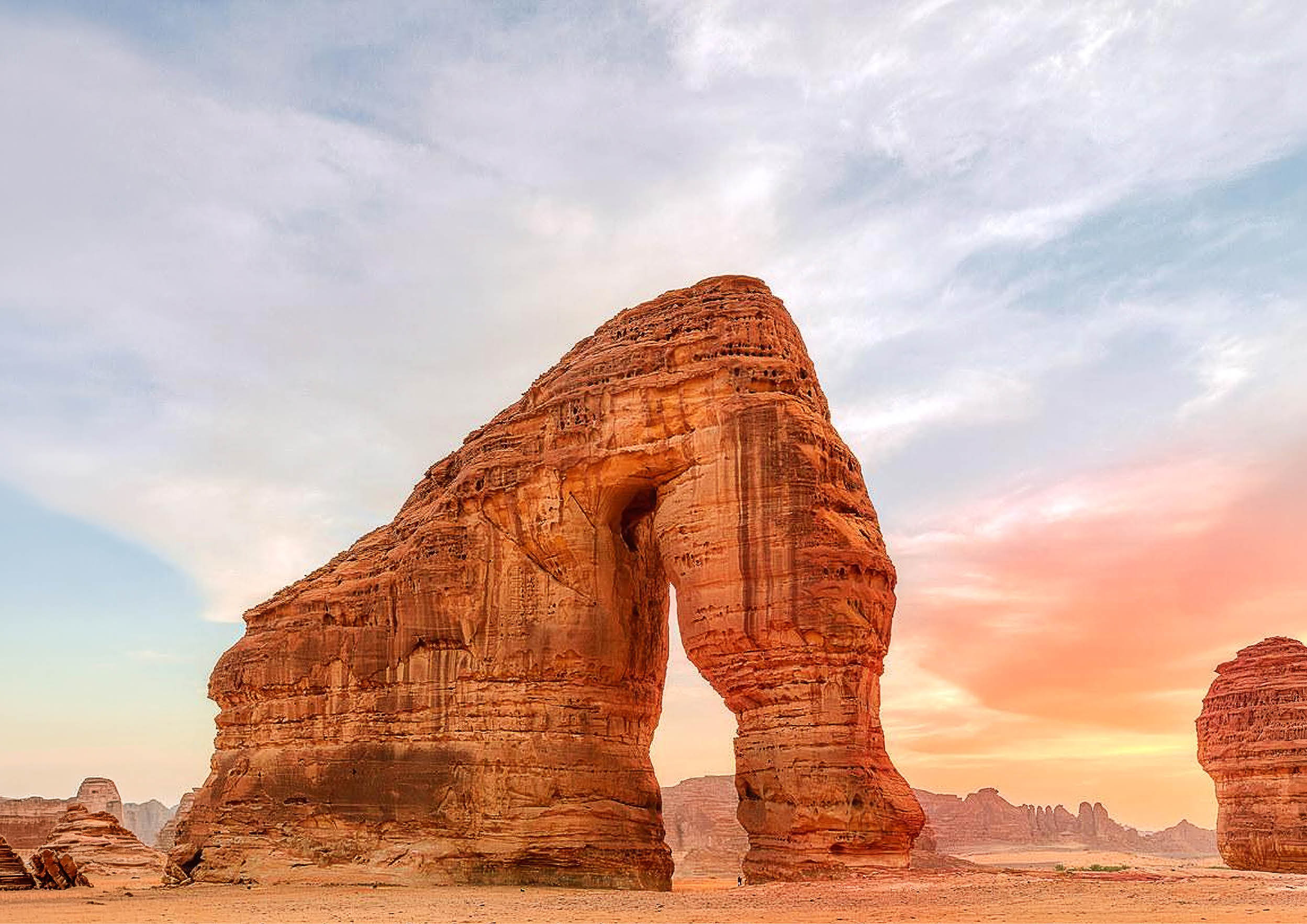صخرة طبيعية ضخمة على شكل فيل في صحراء تحت سماء غائمة مع ألوان شروق أو غروب الشمس. Description generated by AI
