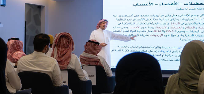 شخص يرتدي ثوبًا تقليديًا يحمل جهاز تحكم عن بعد في يده أمام خلفية بيضاء مع نص عربي جزئي. Description generated by AI