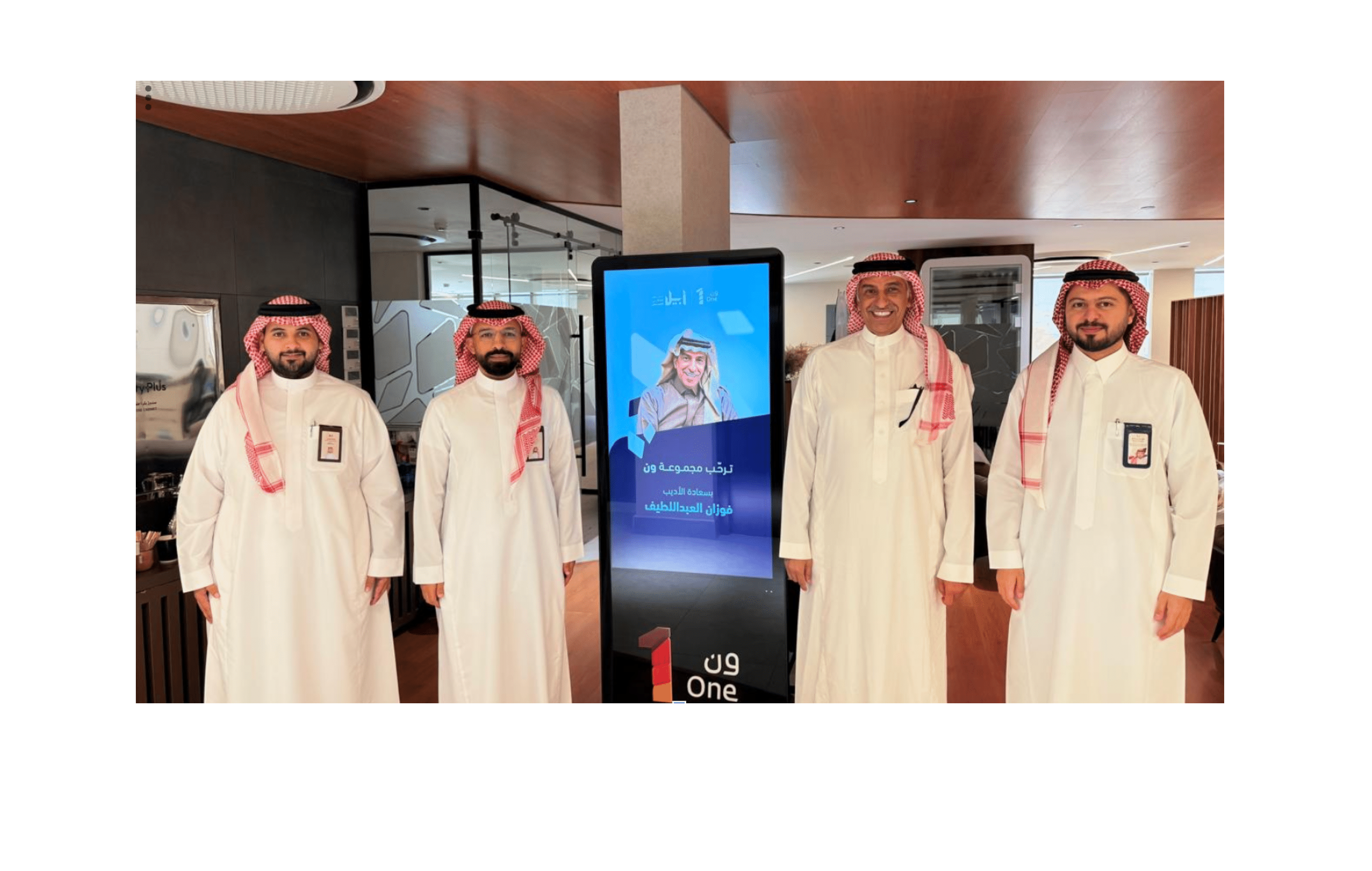 أربعة أشخاص يرتدون الثوب والغترة التقليدية يقفون بجانب شاشة رقمية في مكان داخلي حديث. Description generated by AI