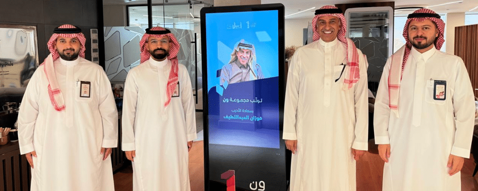 أربعة أشخاص يرتدون الثوب والغترة التقليدية يقفون بجانب شاشة رقمية تعرض صورة ونص باللغة العربية داخل مكان داخلي حديث. Description generated by AI