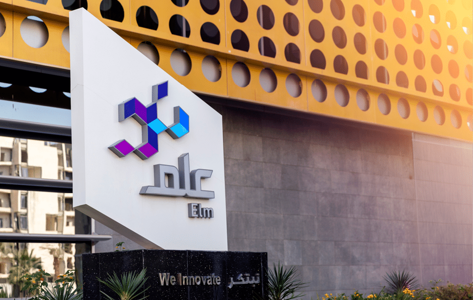 لوحة إعلانية حديثة تحمل شعار شركة علم مع عبارة “We Innovate" مكتوبة بالإنجليزية والعربية، وتقع أمام مبنى حديث مع نباتات في المقدمة. Description generated by AI