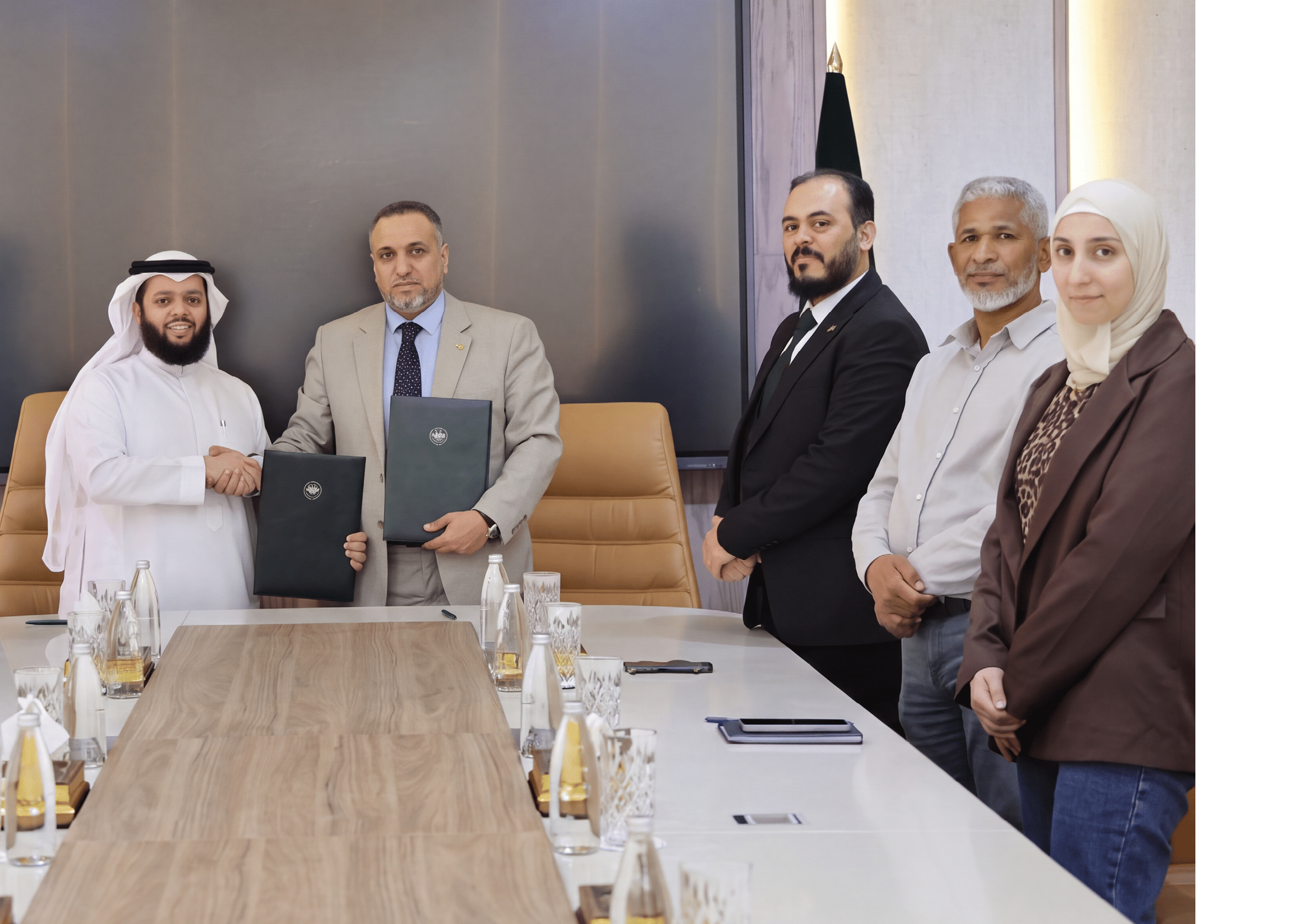 مجموعة من الأشخاص يرتدون ملابس رسمية ويصافحون بعضهم البعض في غرفة اجتماعات حديثة تحتوي على طاولة كبيرة وشاشة عرض خلفهم. Description generated by AI