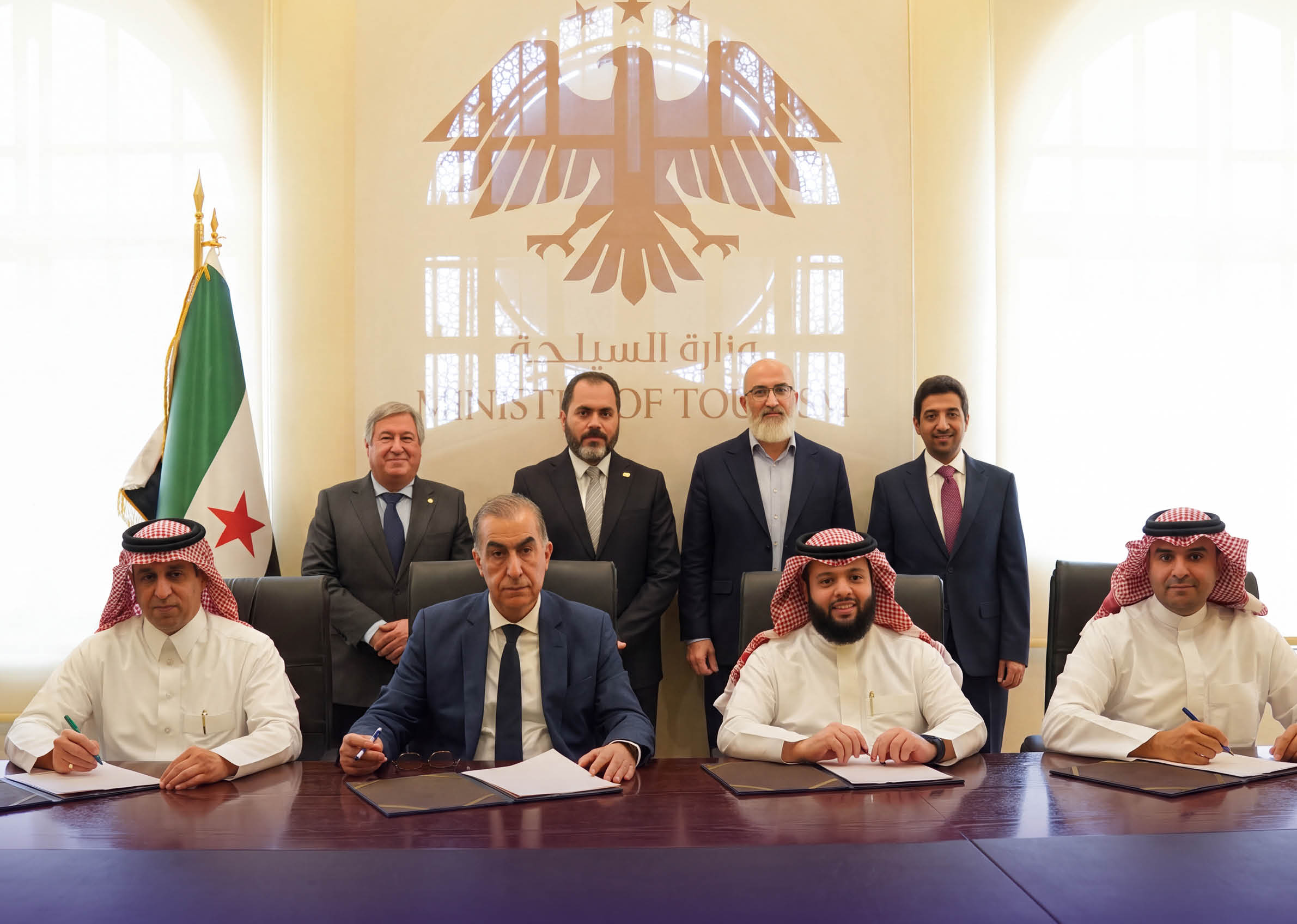 مجموعة من الأشخاص يرتدون ملابس تقليدية ورسمية يجلسون ويوقعون مستندات على طاولة في غرفة رسمية مزينة بعلم وشعار خلفهم. Description generated by AI