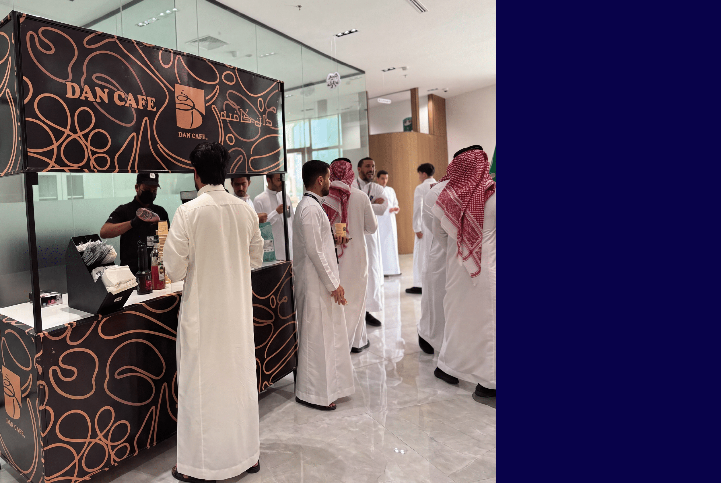 مجموعة من الأشخاص يرتدون الثوب السعودي الأبيض يقفون ويتحدثون بجانب كشك قهوة يحمل شعار دان كافيه في داخل مبنى حديث. Description generated by AI