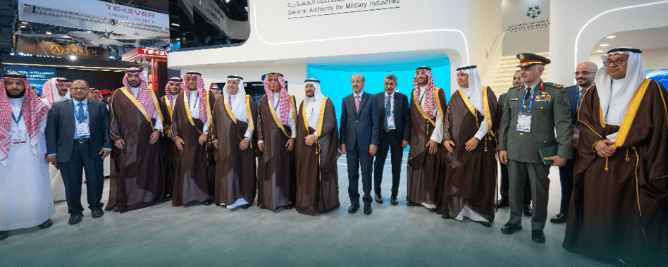 مجموعة من الأشخاص يرتدون ملابس رسمية تقليدية وعسكرية وأزياء عمل يقفون في صف أمام خلفية زرقاء في حدث رسمي. Description generated by AI